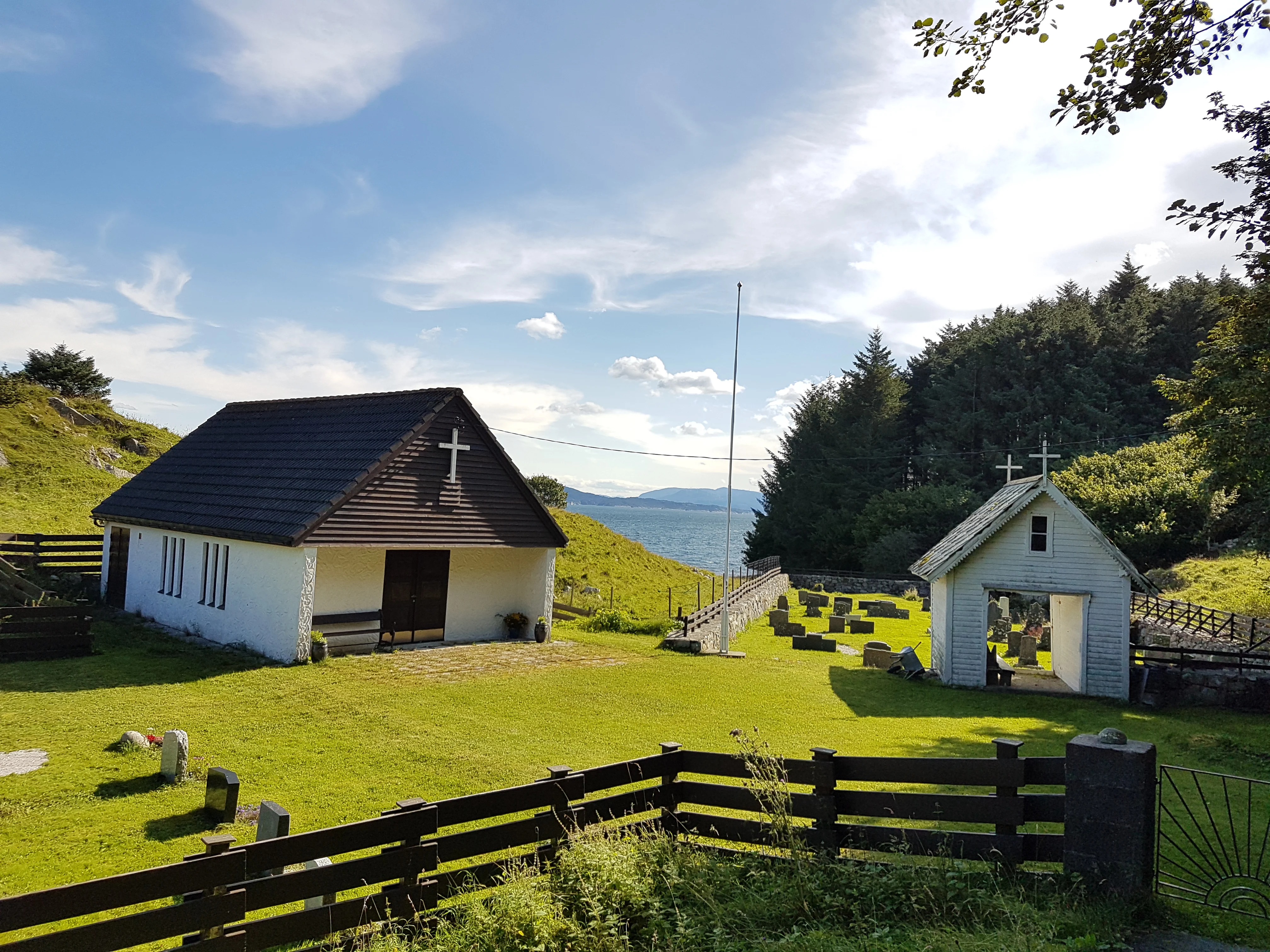 Møkster kapell ligg idyllisk til med utsikt mot havet. Den kvite bygningen med brunt tak er omkransa av grøne marker og gravsteinar, og eit lite kvitt klokketårn står ved sida. Den frodige naturen og den klare himmelen gir ein fredfull atmosfære. Foto.