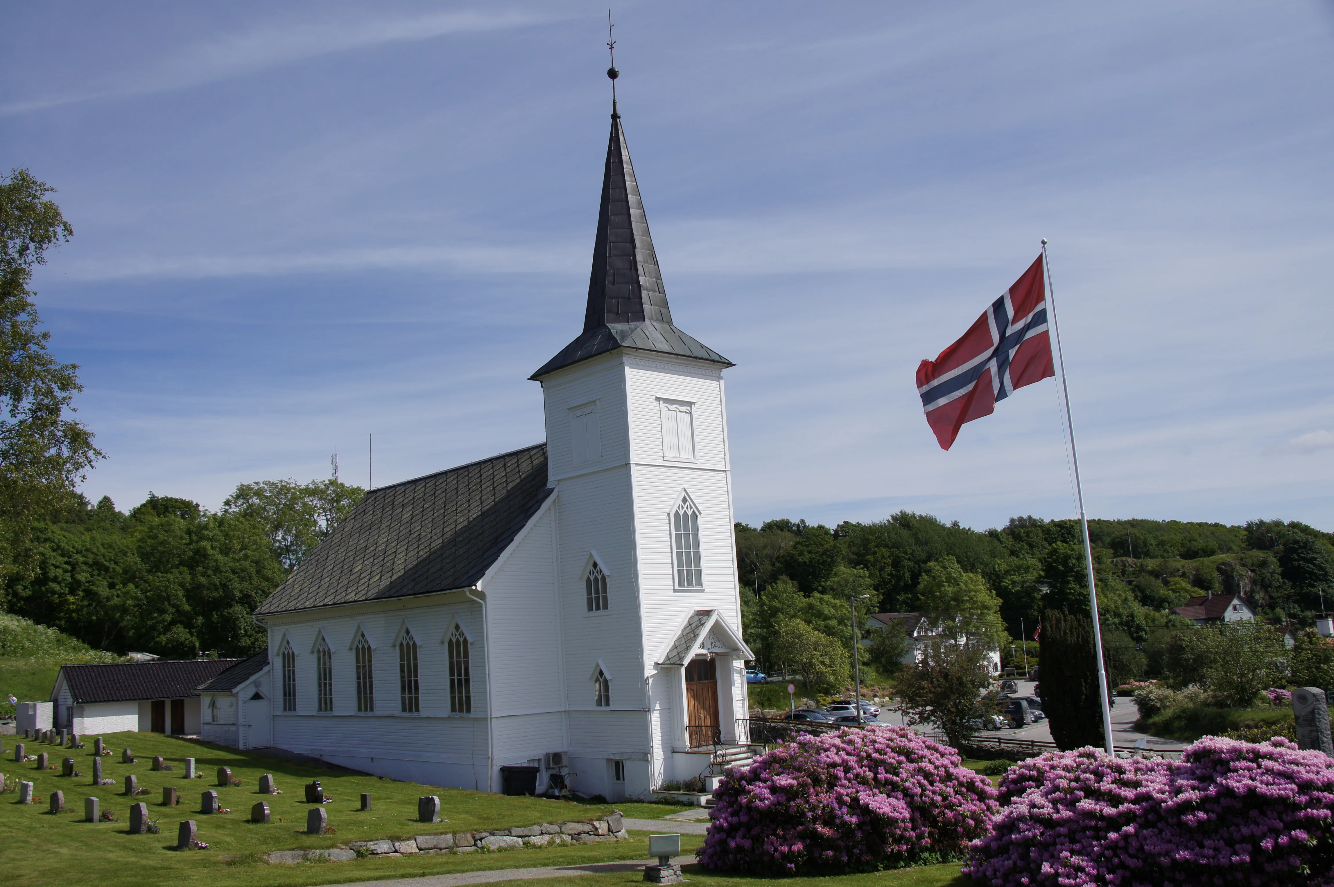 Austevoll kyrkje, ei kvit trekyrkje med høgt spir, omgitt av grøn natur og vakre blomar. Det norske flagget vaier stolt i vinden ved sida av kyrkja. Gravsteinar pryder den velstelte plenen framfor kyrkja. Foto.