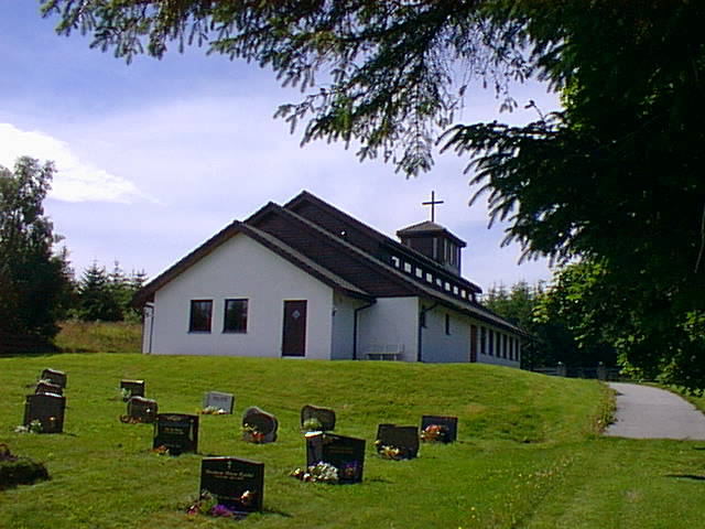 Hundvåkøy kyrkje, ein moderne kvit kyrkjebygning med brunt tak og eit lite klokketårn, ligg vakkert til i grøne omgivnader. Gravsteinar pryder den velstelte plenen framfor kyrkja. Foto.