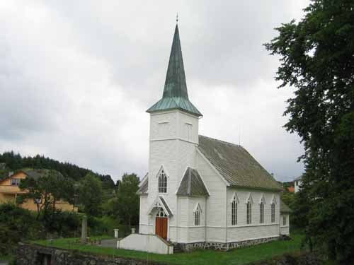 Stolmen kyrkje, ein kvit trekyrkje med grønt tak og eit høgt spir, står stolt i dei frodige omgivnadene. Den tradisjonelle arkitekturen og dei store vindauga gir kyrkja eit ærverdig preg. Foto.