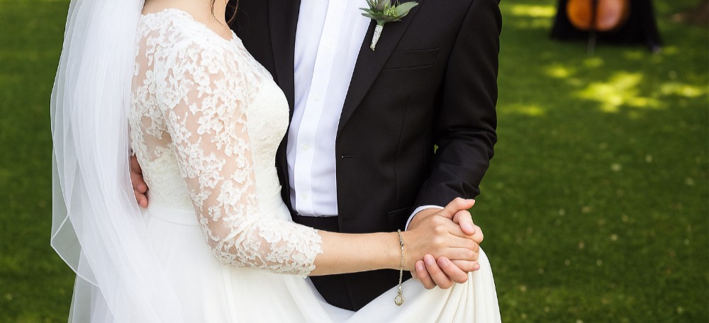 Eit brudepar held hender under bryllaupsseremonien. Brudgommen er kledd i ein mørk dress, medan bruda har på seg ein kvit kjole. Dei har på seg gifteringar, og biletet fokuserer på hendene deira, som symboliserer kjærleik og samhald. Foto.